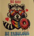 Illustration of a raccoon wearing heart-shaped sunglasses with rainbow colors, holding a donut, and surrounded by hearts with text 'BE GAY BE FABULOUS' on a beige background.